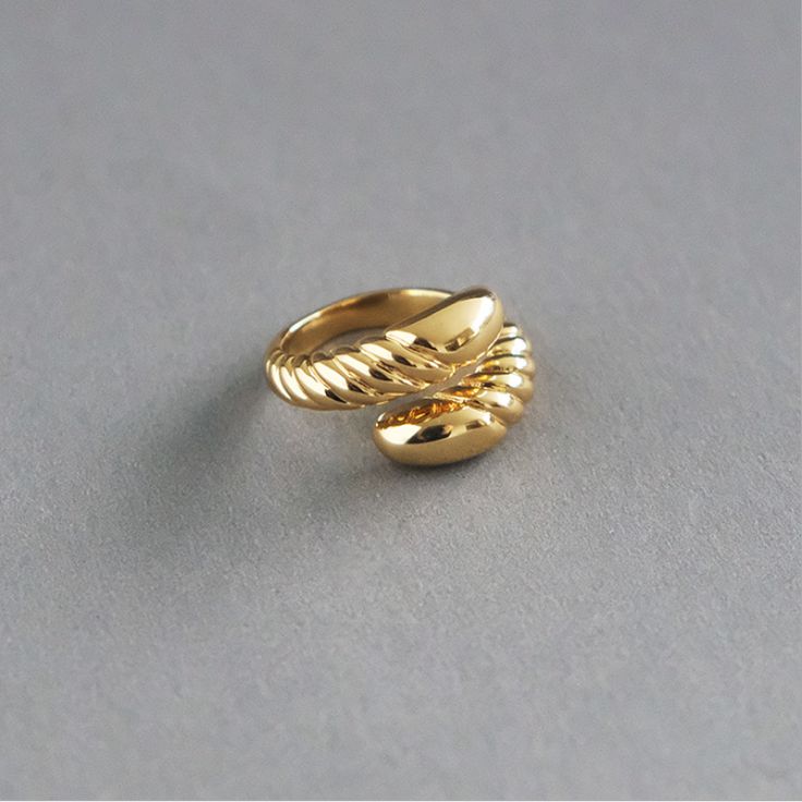 Anel de aço cirúrgico banhado a ouro 18K com design barroco. A peça apresenta uma estrutura robusta com acabamento polido espelhado e detalhes em relevo torcido que criam um contraste sofisticado. O anel está sobre uma superfície cinza lisa, com iluminação suave destacando seu brilho e textura.