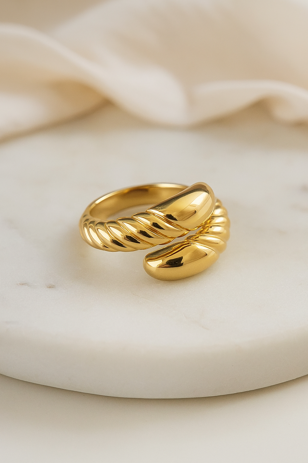 Anel de aço cirúrgico banhado a ouro 18K com design barroco. A peça apresenta uma estrutura robusta com acabamento polido espelhado e detalhes em relevo torcido que criam um contraste sofisticado. O anel está sobre uma superfície cinza lisa, com iluminação suave destacando seu brilho e textura.