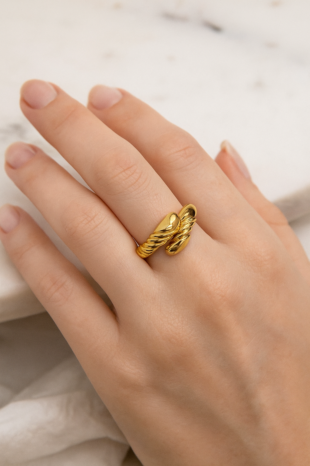 Anel de aço cirúrgico banhado a ouro 18K com design barroco. A peça apresenta uma estrutura robusta com acabamento polido espelhado e detalhes em relevo torcido que criam um contraste sofisticado. O anel está sobre uma superfície cinza lisa, com iluminação suave destacando seu brilho e textura.