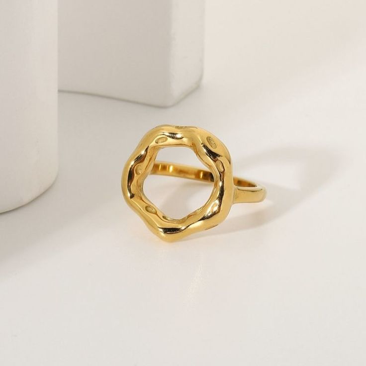 Anel dourado com design escultural e aberto, formado por um aro grosso e irregular em formato circular orgânico, com acabamento polido e levemente ondulado, evocando a imagem de um eclipse. A peça tem superfície espelhada e textura fluida, destacando-se em fundo claro minimalista.