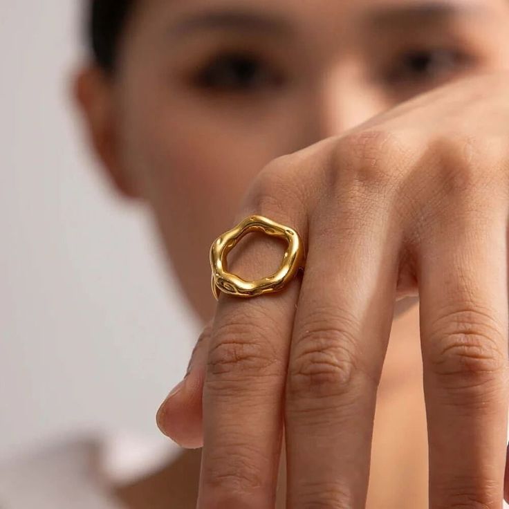 Anel dourado com design escultural e aberto, formado por um aro grosso e irregular em formato circular orgânico, com acabamento polido e levemente ondulado, evocando a imagem de um eclipse. A peça tem superfície espelhada e textura fluida, destacando-se em fundo claro minimalista.
