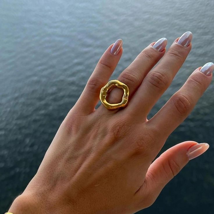 Anel dourado com design escultural e aberto, formado por um aro grosso e irregular em formato circular orgânico, com acabamento polido e levemente ondulado, evocando a imagem de um eclipse. A peça tem superfície espelhada e textura fluida, destacando-se em fundo claro minimalista.
