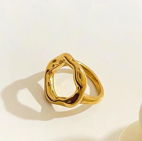 Anel dourado com design escultural e aberto, formado por um aro grosso e irregular em formato circular orgânico, com acabamento polido e levemente ondulado, evocando a imagem de um eclipse. A peça tem superfície espelhada e textura fluida, destacando-se em fundo claro minimalista.