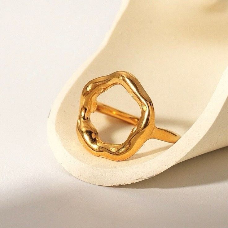 Anel dourado com design escultural e aberto, formado por um aro grosso e irregular em formato circular orgânico, com acabamento polido e levemente ondulado, evocando a imagem de um eclipse. A peça tem superfície espelhada e textura fluida, destacando-se em fundo claro minimalista.