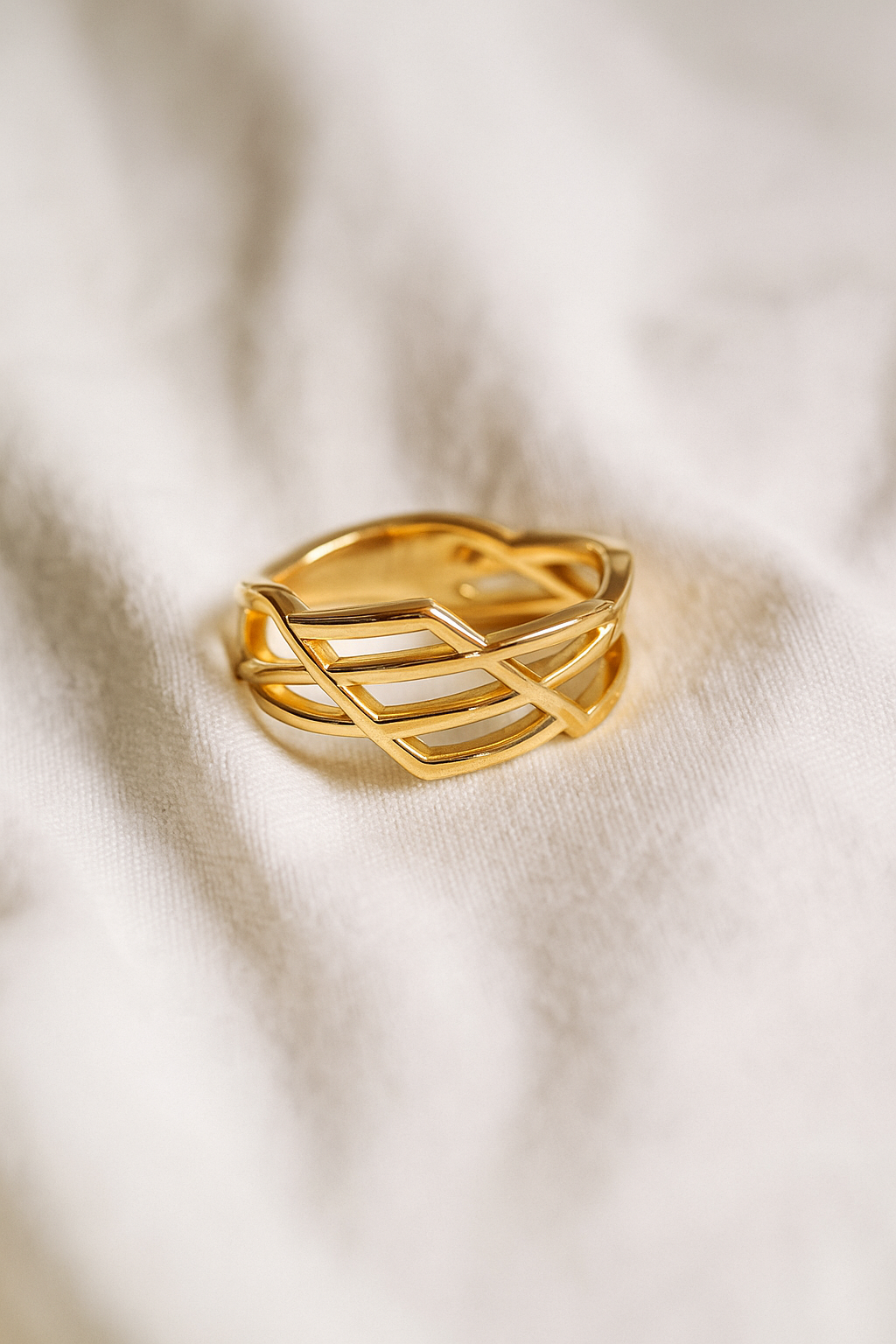 Anel dourado de aço cirúrgico banhado a ouro 18K, com design geométrico entrelaçado, formado por fios finos que se cruzam, fotografado sobre superfície neutra clara.