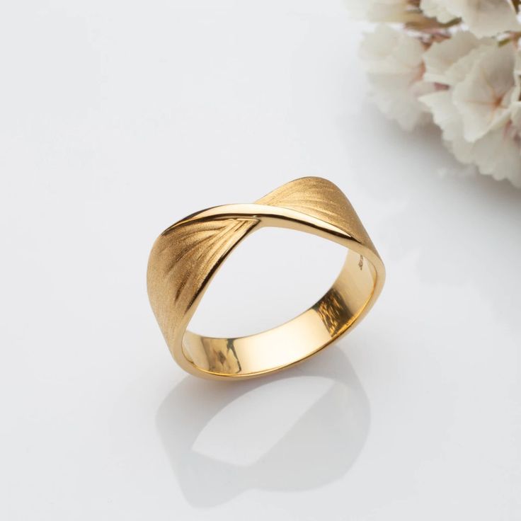 Anel dourado com design moderno exibido sobre superfície branca brilhante ao lado de flores brancas. O anel possui um leve torcimento no centro, com acabamento fosco na parte externa e polido espelhado na parte interna. A torção forma linhas gravadas que irradiam suavemente para as laterais, criando um efeito elegante e minimalista. A iluminação é suave, com sombras discretas realçando os detalhes do anel.