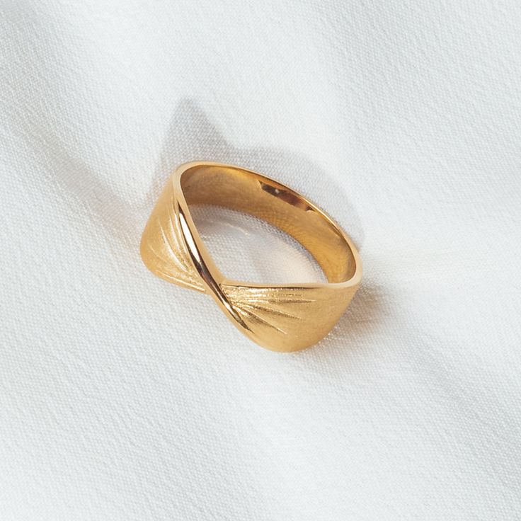 Anel dourado com design moderno exibido sobre superfície branca brilhante ao lado de flores brancas. O anel possui um leve torcimento no centro, com acabamento fosco na parte externa e polido espelhado na parte interna. A torção forma linhas gravadas que irradiam suavemente para as laterais, criando um efeito elegante e minimalista. A iluminação é suave, com sombras discretas realçando os detalhes do anel.