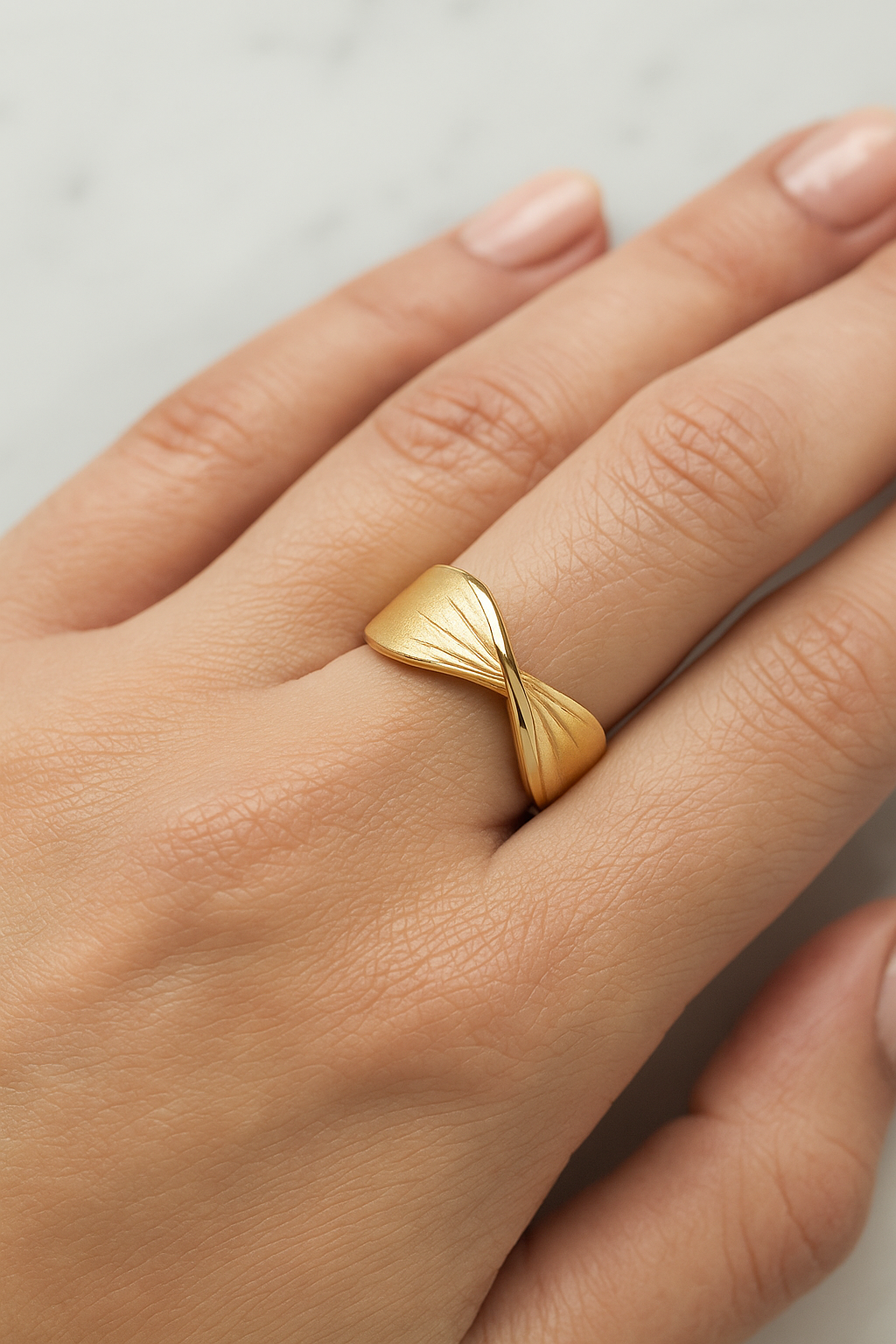 Anel dourado com design moderno exibido sobre superfície branca brilhante ao lado de flores brancas. O anel possui um leve torcimento no centro, com acabamento fosco na parte externa e polido espelhado na parte interna. A torção forma linhas gravadas que irradiam suavemente para as laterais, criando um efeito elegante e minimalista. A iluminação é suave, com sombras discretas realçando os detalhes do anel.