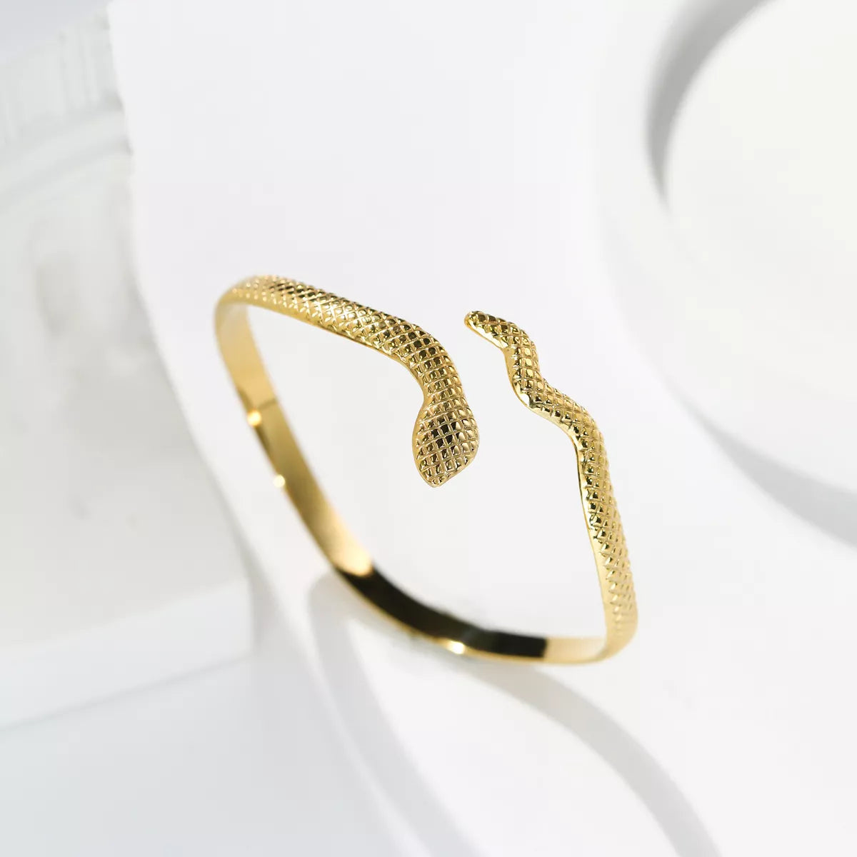 Pulseira rígida dourada com design de serpente estilizada, acabamento texturizado e abertura frontal em formato de cabeça e cauda de cobra, sobre fundo branco.