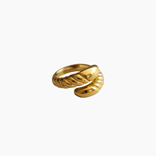 Anel de aço cirúrgico banhado a ouro 18K com design barroco. A peça apresenta uma estrutura robusta com acabamento polido espelhado e detalhes em relevo torcido que criam um contraste sofisticado. O anel está sobre uma superfície cinza lisa, com iluminação suave destacando seu brilho e textura.