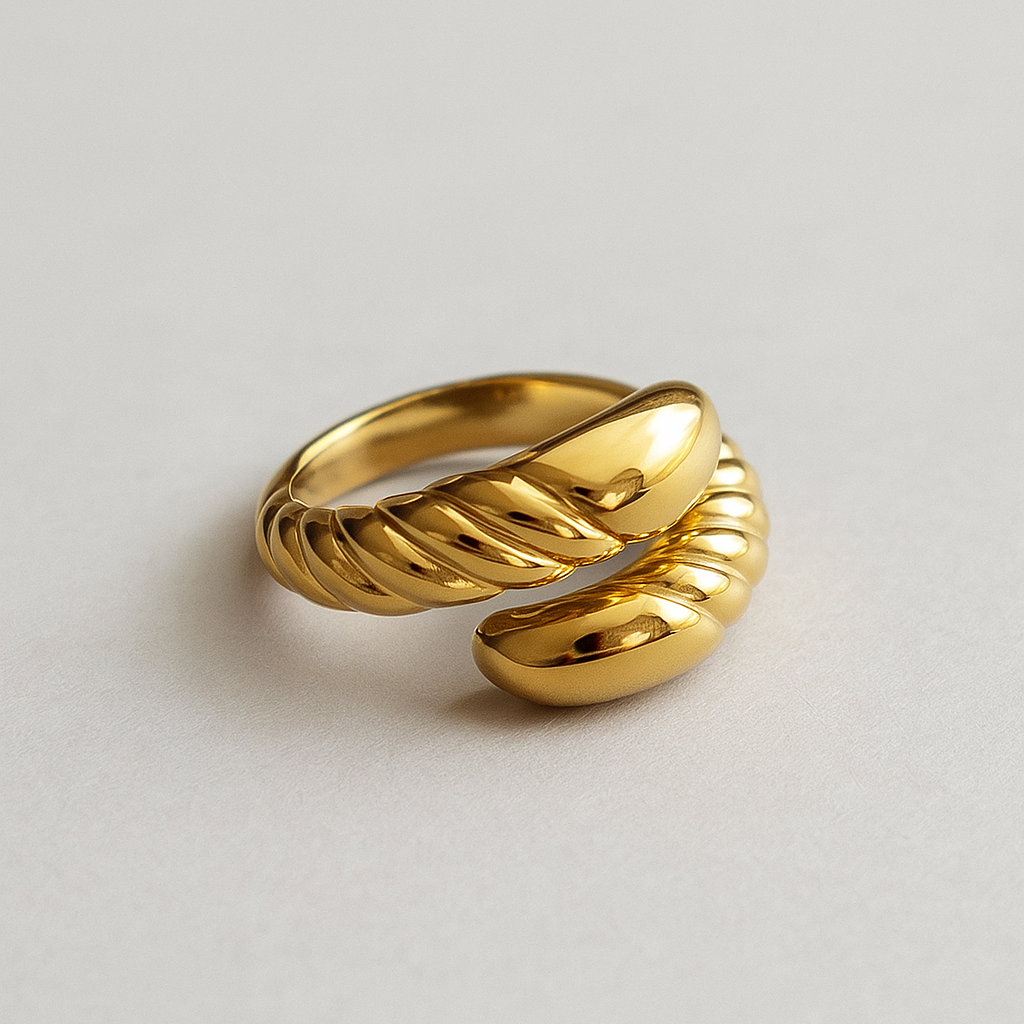 Anel de aço cirúrgico banhado a ouro 18K com design barroco. A peça apresenta uma estrutura robusta com acabamento polido espelhado e detalhes em relevo torcido que criam um contraste sofisticado. O anel está sobre uma superfície cinza lisa, com iluminação suave destacando seu brilho e textura.