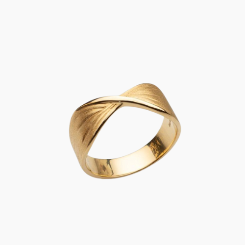 Anel dourado com design moderno exibido sobre superfície branca brilhante ao lado de flores brancas. O anel possui um leve torcimento no centro, com acabamento fosco na parte externa e polido espelhado na parte interna. A torção forma linhas gravadas que irradiam suavemente para as laterais, criando um efeito elegante e minimalista. A iluminação é suave, com sombras discretas realçando os detalhes do anel.
