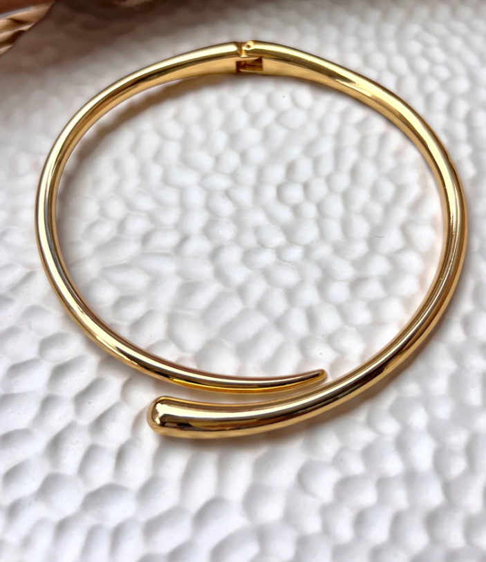Colar rígido banhado a ouro 18K, com formato de meia-lua aberta e acabamento polido espelhado, posicionado ao redor do pescoço de uma modelo com top preto e fundo bege neutro.